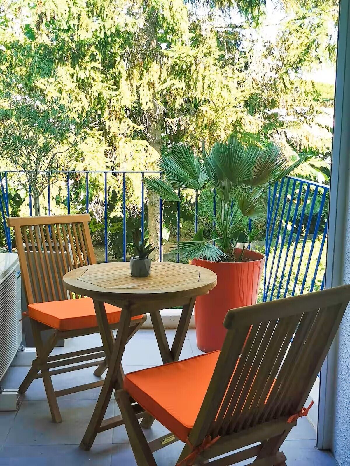 A balcony with a round wooden table, two wooden chairs with orange cushions, and a potted plant.