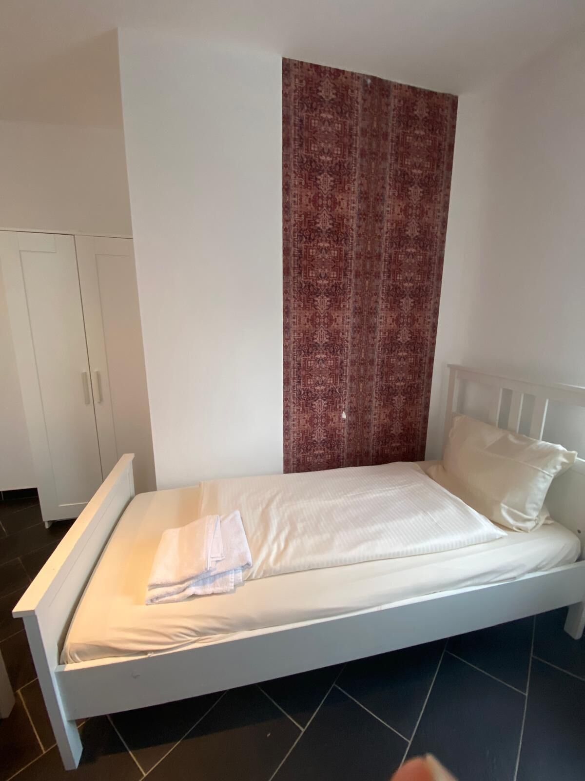 Ein Zimmer mit einem Einzelbett, einem roten Vorhang mit Muster, weißen Wänden und einem weißen Kleiderschrank.