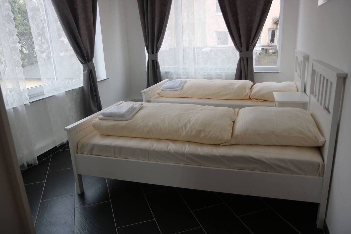 Ein Doppelbett mit weißer Bettwäsche und einem gefalteten Handtuch, ein Fenster mit Vorhängen und ein gefliester Boden.