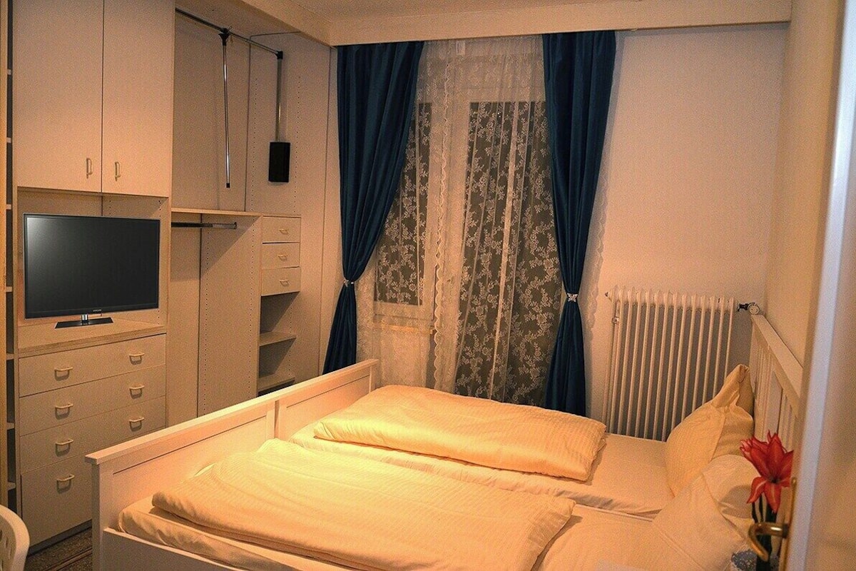 Ein Schlafzimmer mit Bett, Fernseher, Kommode und einem Fenster mit Vorhängen.