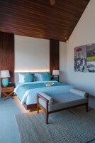 A bedroom with a wooden ceiling, a large bed with a headboard, two bedside tables with lamps, and a wall with two framed pictures.