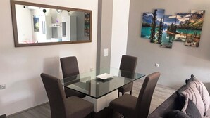 Un salon moderne avec une table à manger au plateau en verre, quatre chaises foncées, un canapé gris et un grand miroir accroché au mur.