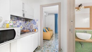 Een keuken met een magnetron, een spoelbak en een decoratieve spatwand. In de aangrenzende woonkamer staat een bank met een blauw gordijn.