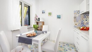 Een compacte keuken met witte kasten, een eettafel met stoelen, een klein eilandje met een plant en een betegelde spatwand.