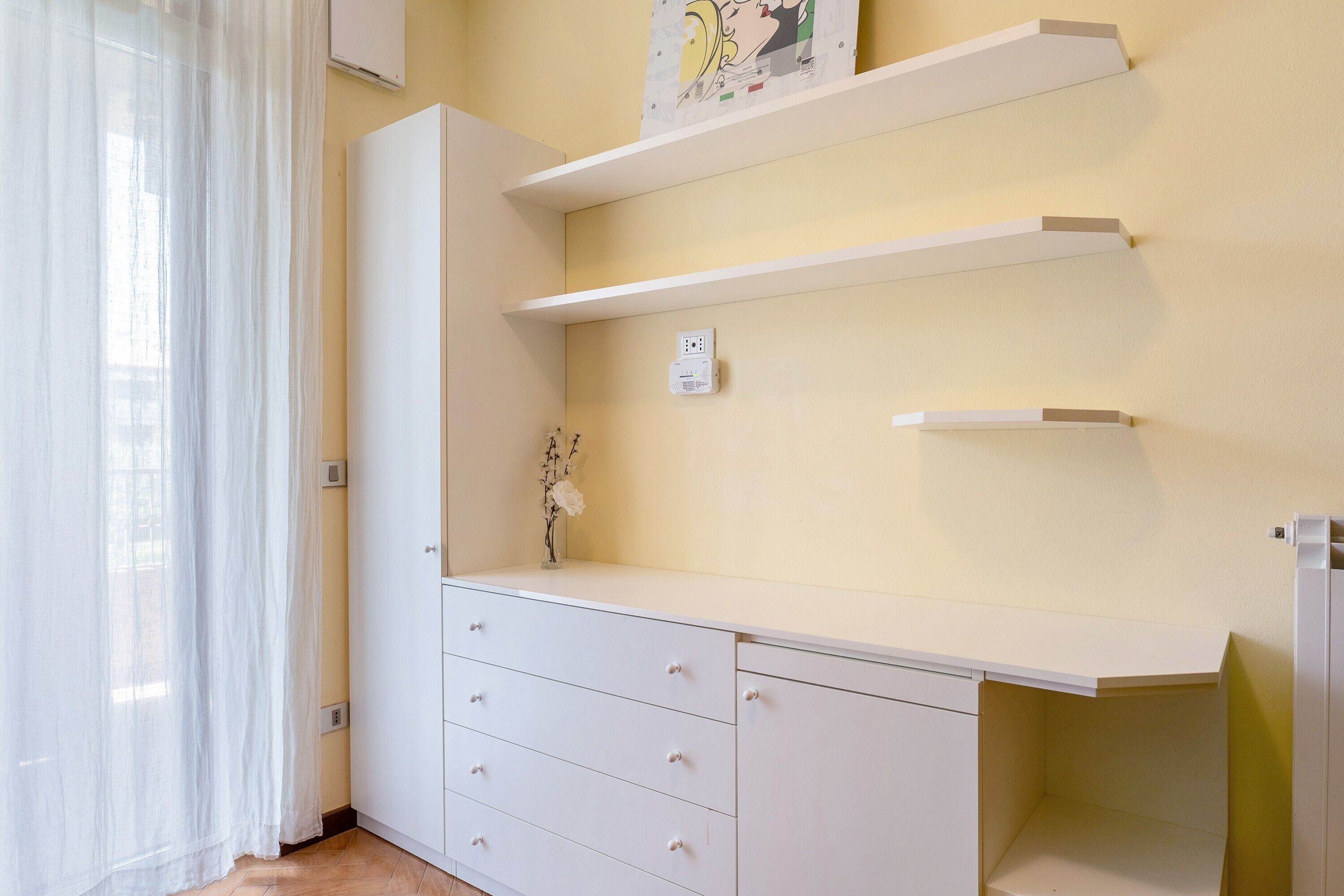 A room with a white wardrobe, a desk with drawers, a white shelf unit, and a wall-mounted electrical outlet.