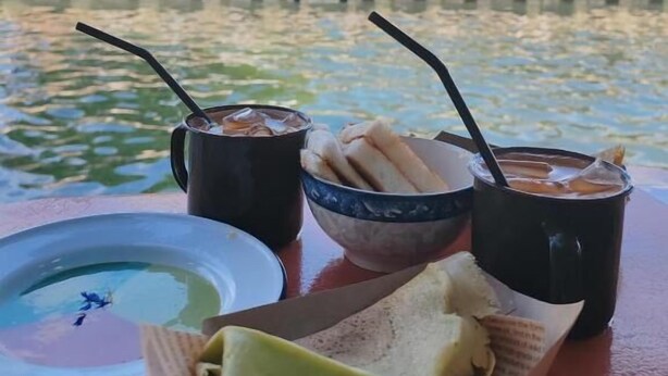 A table with a plate, a bowl of bread, and two cups of a beverage, set against a backdrop of a water body and buildings. - Heeren Street Hotel at Jonker Walk Melaka (Malacca City)