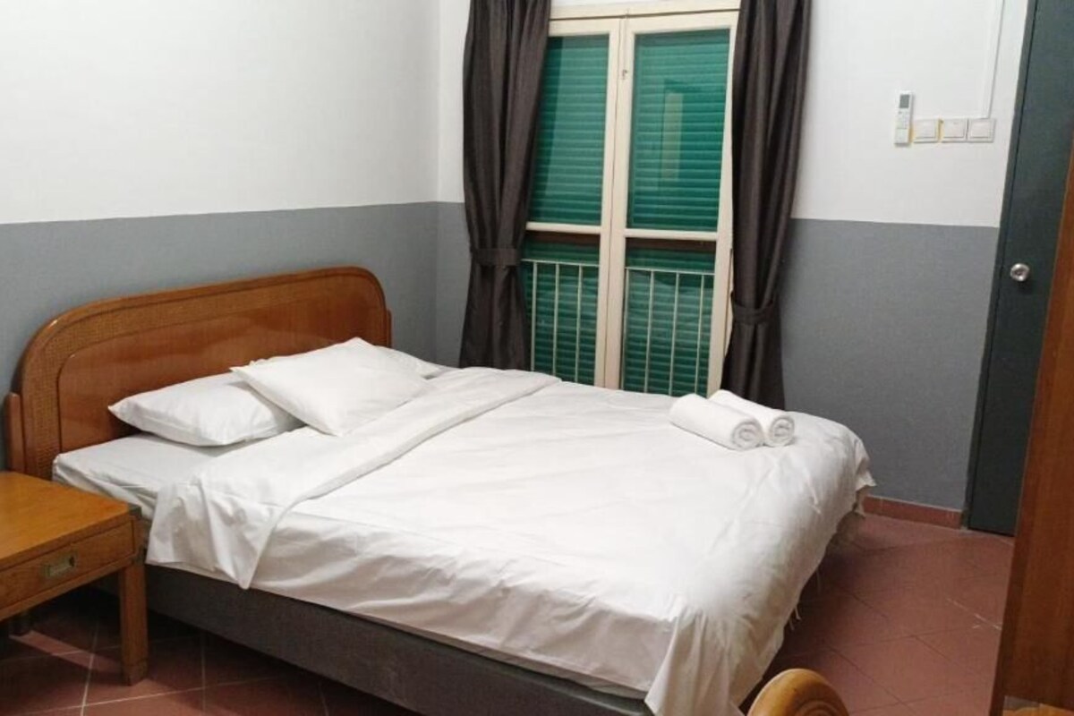 Una cama individual con ropa blanca, una mesita de noche de madera y una ventana con contraventanas verdes.