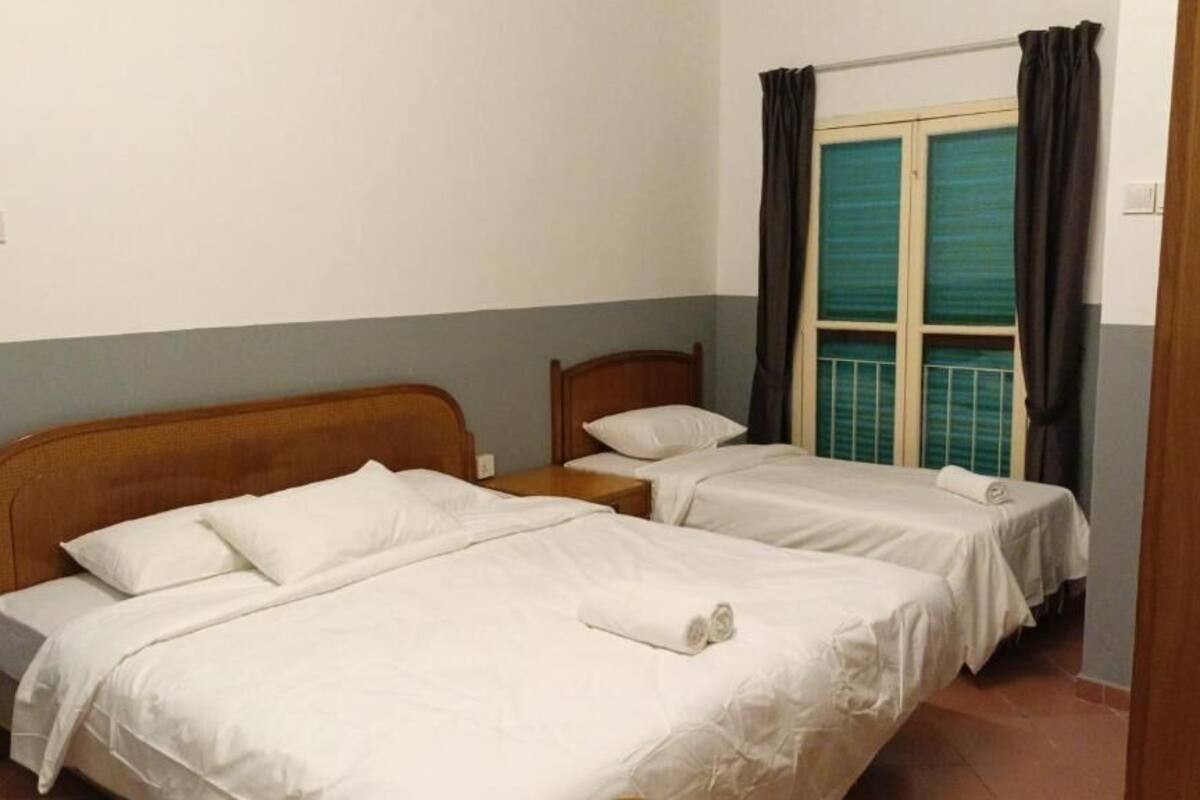Une chambre avec deux lits simples, une tête de lit en bois, une fenêtre avec des rideaux et un sol carrelé.