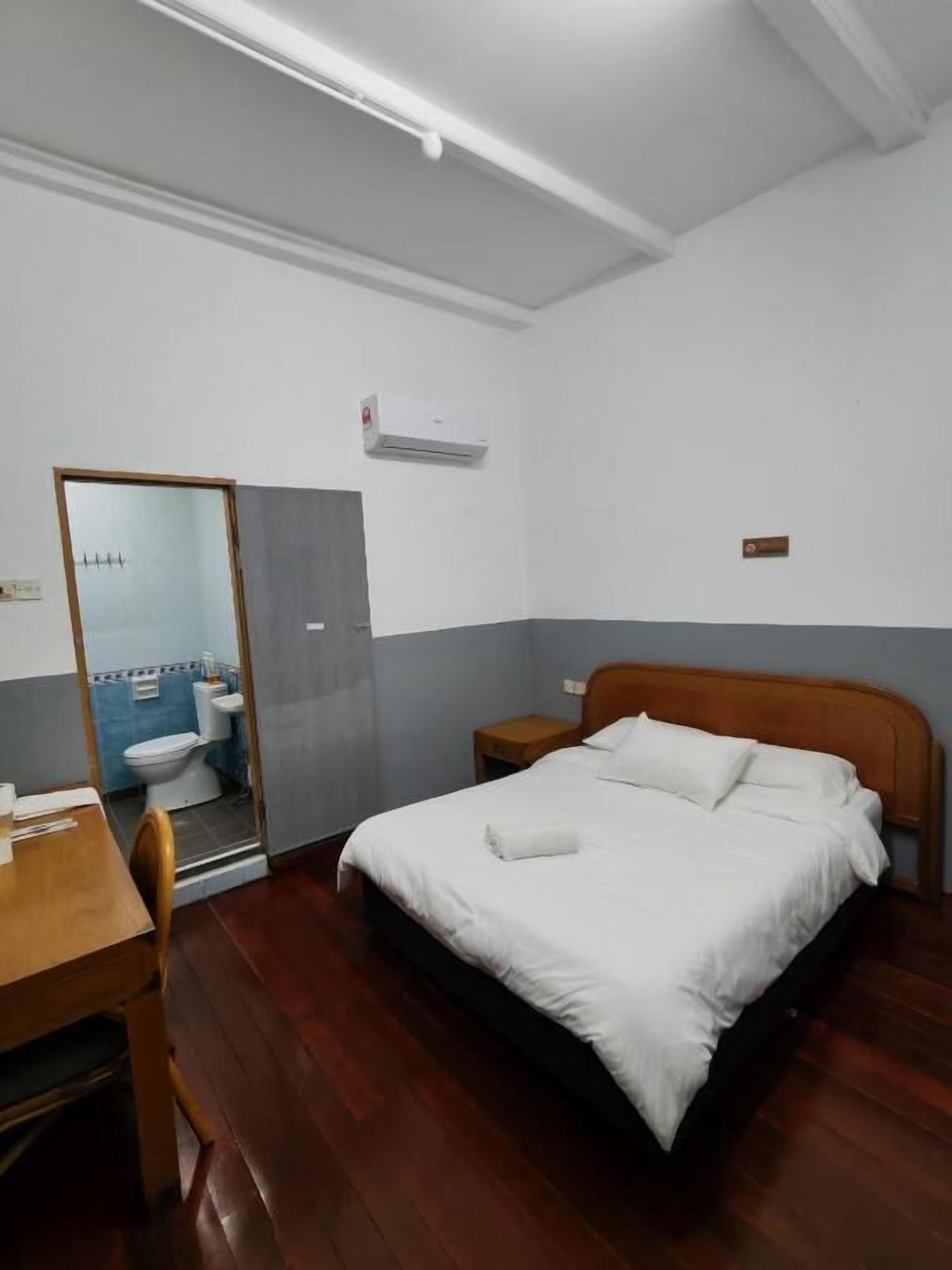 A bedroom with a wooden bed, a bedside table, a desk with a chair, and a bathroom visible through an open door.