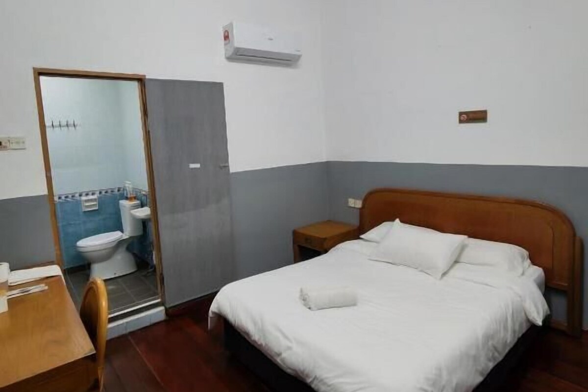 Un dormitorio con cama de madera, una mesita de noche, un escritorio con silla, y un baño visible a través de una puerta abierta.