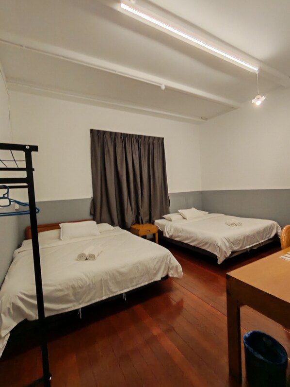 Habitación de hotel con dos camas, una mesa de madera, una silla pequeña, un escritorio, una lámpara de escritorio, un perchero y un cubo de basura azul.
