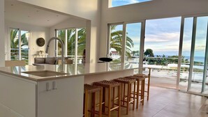 A modern kitchen with a large island, a dining area with bar stools, and a view of the beach and ocean through large windows. - Villa Palmeras - Modern luxury with breathtaking sea view (Caleta de Vélez)