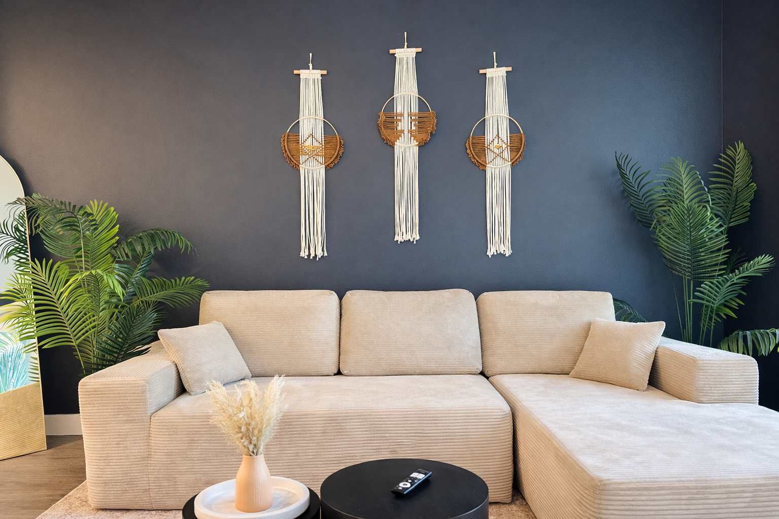 Een moderne woonkamer met een hoekbank, een ronde salontafel en drie macramé wanddecoraties.