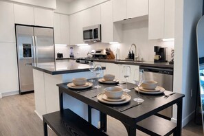 Une cuisine moderne avec des meubles blancs, un réfrigérateur en acier inoxydable, un four à micro-ondes et un coin repas agrémenté d’une table en bois foncé, mise pour quatre personnes.