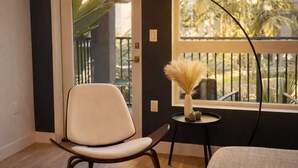 A room with a chair, a small table with a vase, and a view of a balcony with plants. - Designer Apartment w/ Pool & Gym 10 Min to Disney (Santa Ana)