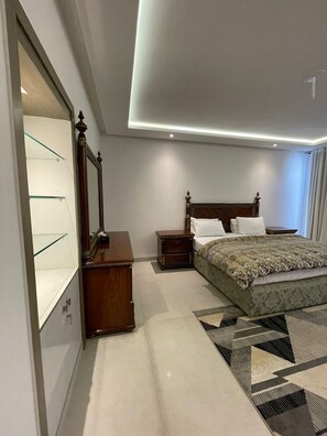 A modern bedroom with a large bed, a wooden nightstand, a dresser with glass doors, and a ceiling light fixture.