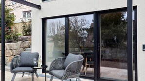A modern outdoor seating area with two grey chairs and a small table, a glass door leading to a patio, and a stone wall. - Rosenhof with whirlpool | Grüne Tanne | seeglueck (Konstanz)