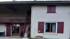 Una casa de dos pisos con camino de grava, una escalera de madera y un balcón con contraventanas rojas.