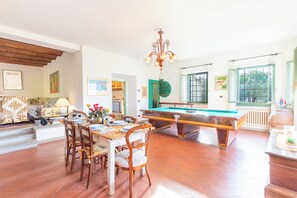 Un vaste espace de vie comprenant une table de billard, une table à manger et des chaises. Les murs sont ornés de tableaux encadrés et un lustre est suspendu au plafond.
