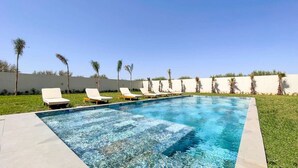 Une piscine rectangulaire à l’eau d’un bleu cristallin, entourée de chaises longues blanches et de palmiers.