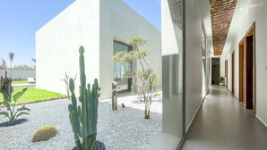 Un bâtiment moderne au design épuré et minimaliste, doté d’une cour en gravier agrémentée de cactus et de petits arbres, ainsi que d’un long couloir étroit avec des portes en bois et un plafond orné d’un motif géométrique.
