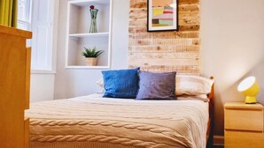Ein ordentlich eingerichtetes Schlafzimmer mit einem Bett, einem Nachttisch mit Lampe, einem Regal mit Pflanze und einem gerahmten Kunstwerk an der Wand.