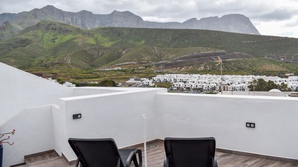Un balcón con dos sillas y una mesa, con vistas a un pueblo de edificios blancos y colinas verdes al fondo.