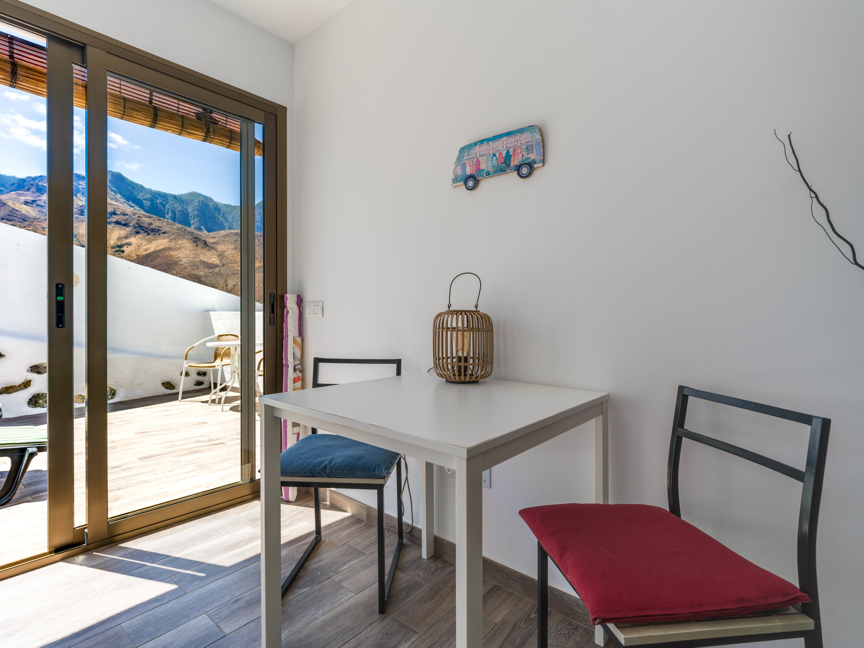 Quarto com uma mesa branca, uma cadeira vermelha, um candeeiro de vime e vista para montanhas através de uma porta de vidro.