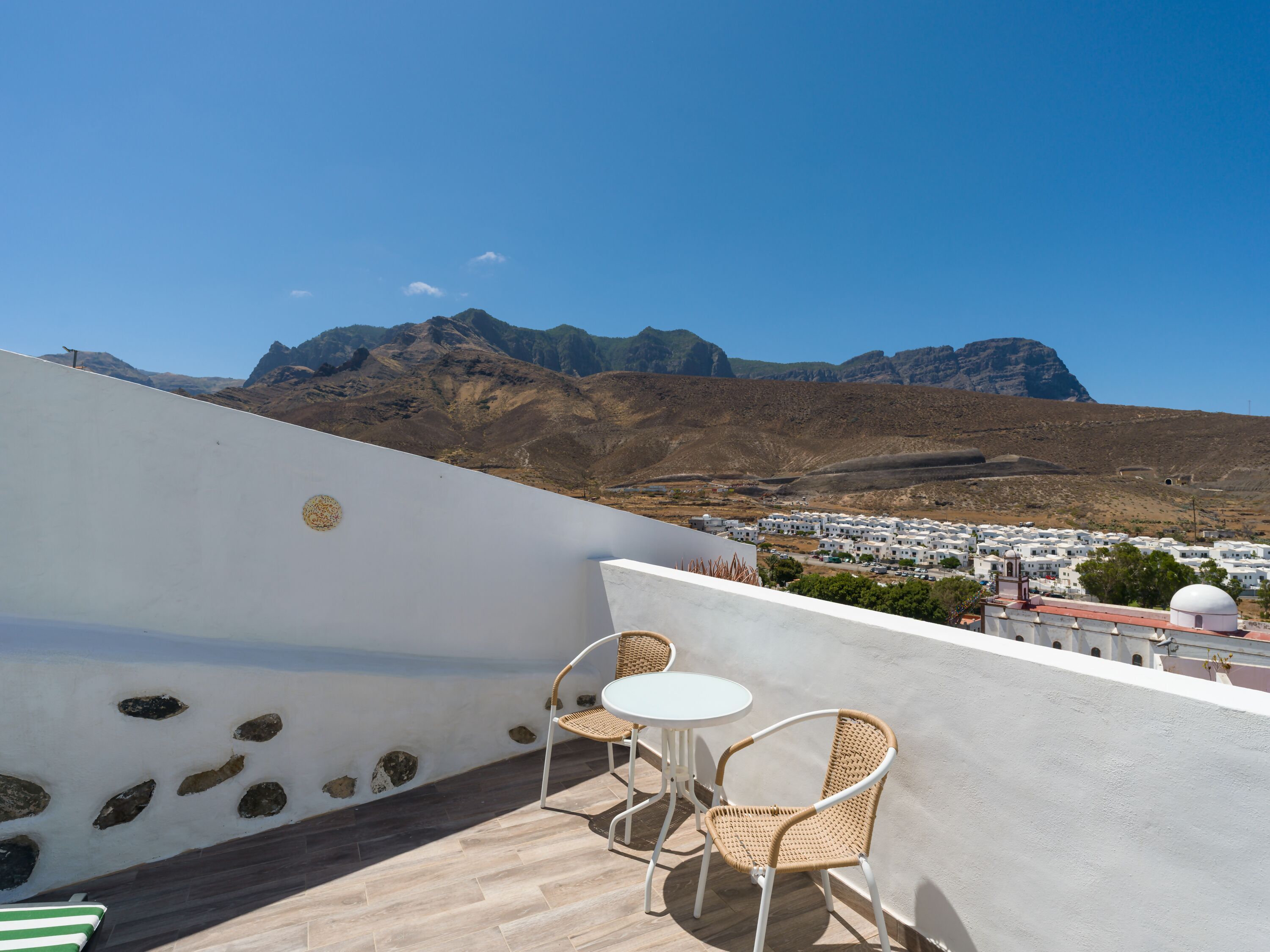 A terrace with two wicker chairs and a small table, overlooking a town and mountains.