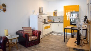 Une cuisine compacte avec des meubles blancs et jaunes, un réfrigérateur blanc et un four à micro-ondes. On y trouve un coin repas avec une petite table et une chaise, ainsi qu'un canapé avec des coussins à rayures.