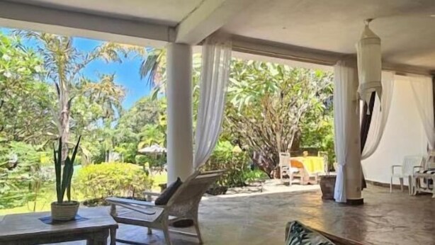 Um espaço amplo e arejado com vista para vegetação exuberante. Há um banco de madeira, uma área de estar acolchoada e uma mesa com um vaso de planta.