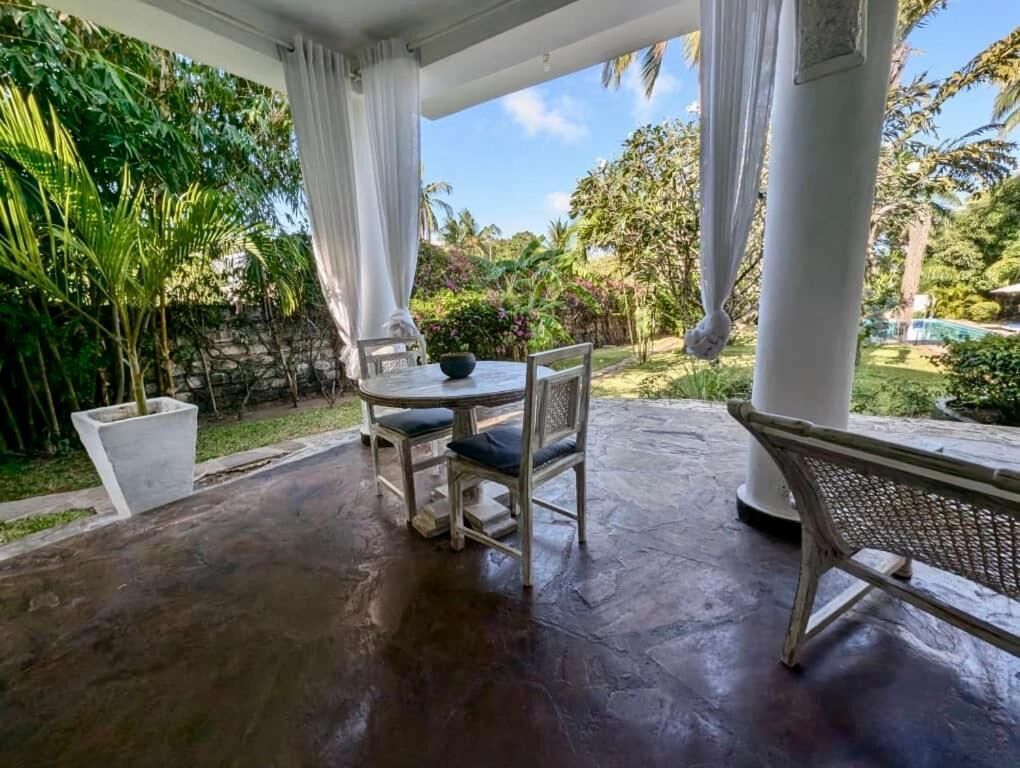 A covered patio with a table and chairs, surrounded by lush greenery and a stone wall.