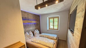 Ein Schlafzimmer mit hölzernem Kopfteil, zwei Betten mit gemusterten Bettbezügen, einem kleinen Fenster mit Ausblick und einem Fernseher auf einem Holzgestell.