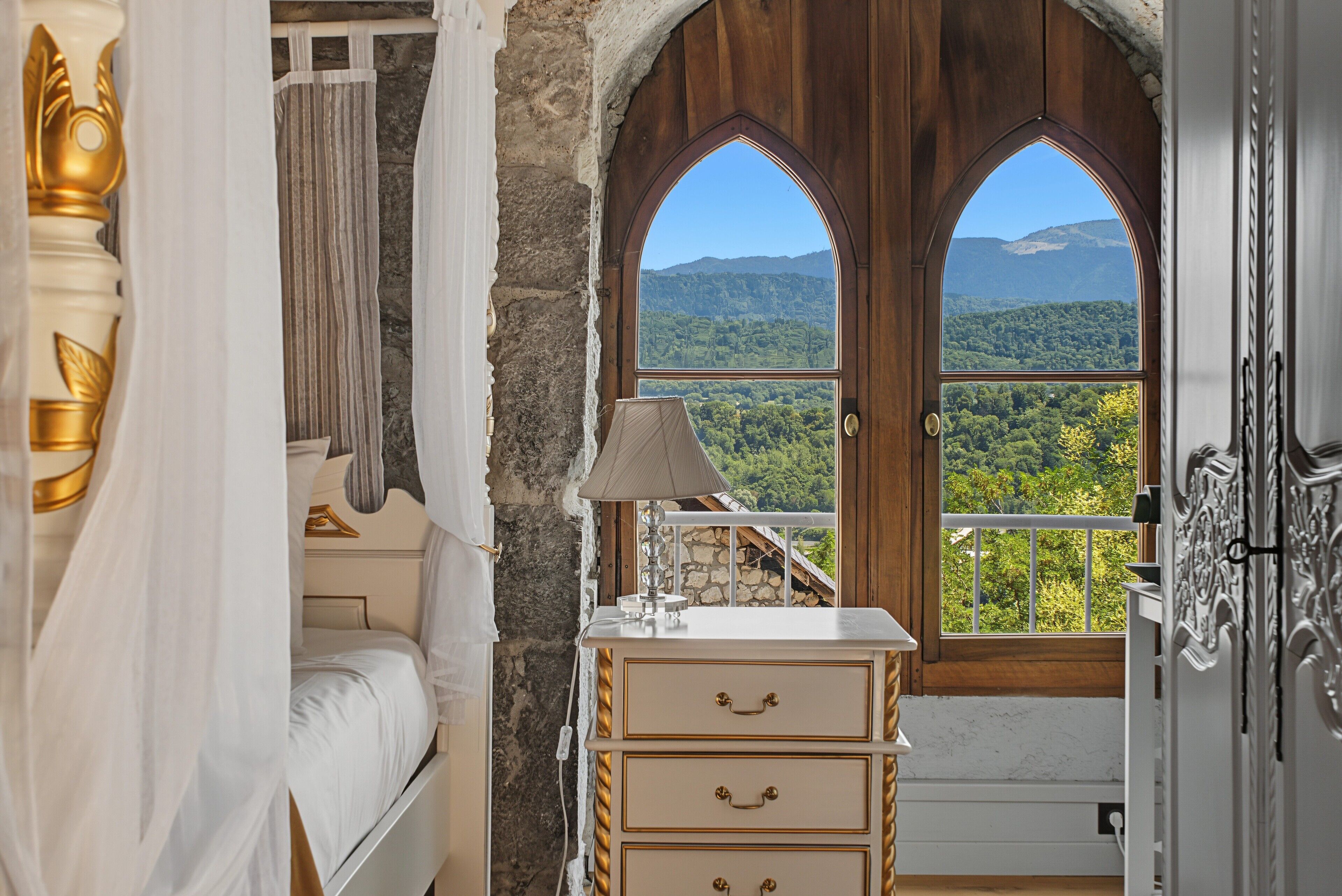 A bedroom with a bed, a bedside lamp, a dresser, and a view of a mountainous landscape through a large arched window.