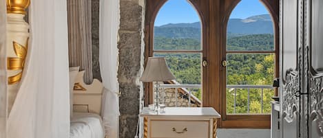 A bedroom with a bed, a bedside lamp, a dresser, and a view of a mountainous landscape through a large arched window.