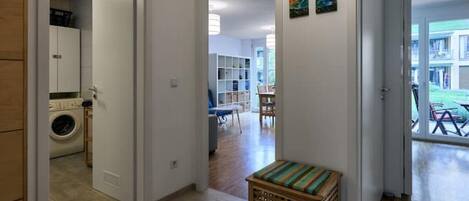 A hallway leading to a living area with a laundry room on the left, a small seating area with a bench, and a view of the outside balcony.