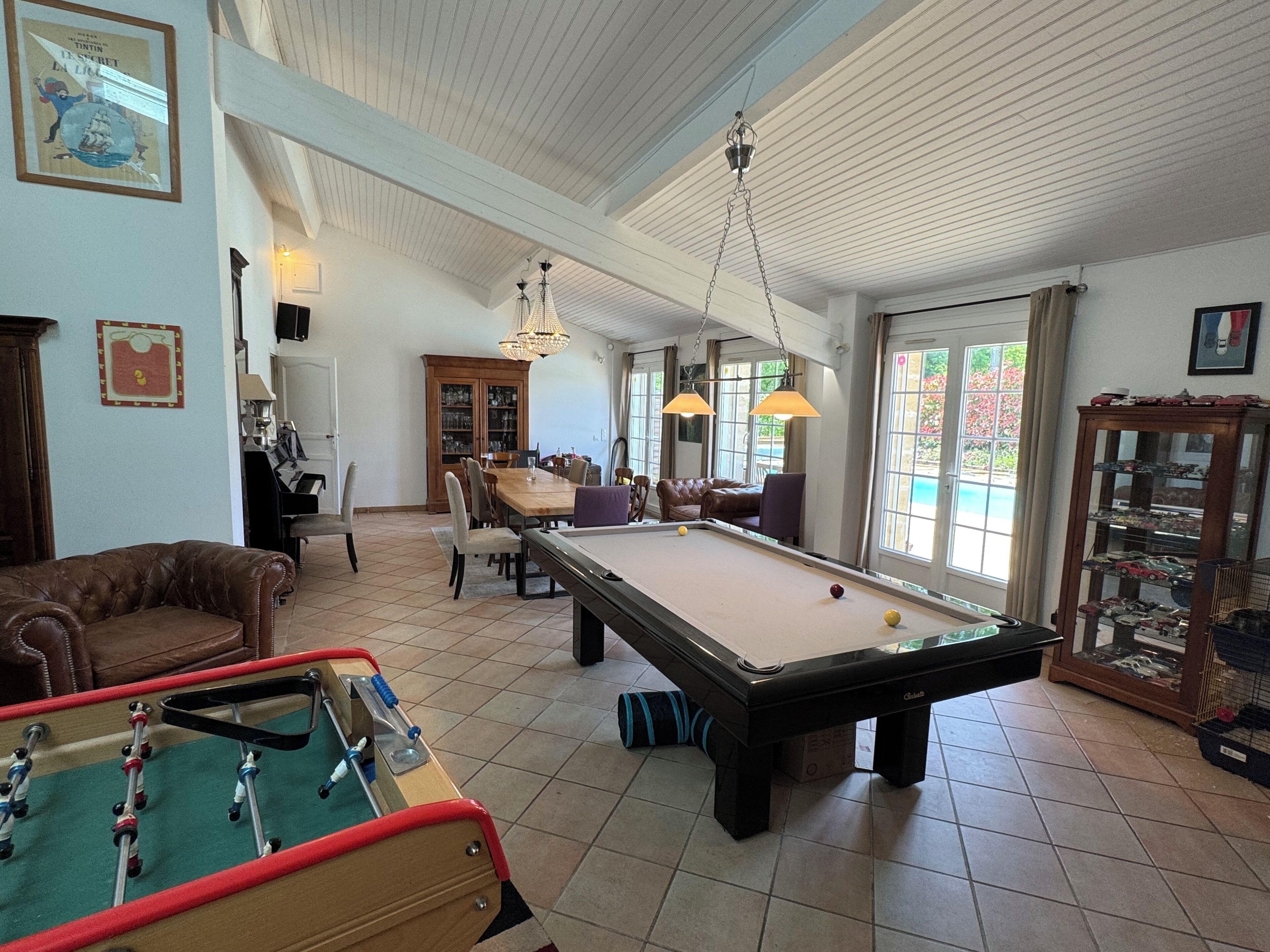 A spacious room with a pool table, a foosball table, and a billiards table. There is a dining area with a wooden cabinet and chairs.