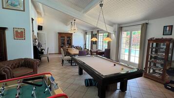 A spacious room with a pool table, a foosball table, and a billiards table. There is a dining area with a wooden cabinet and chairs.