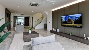 A modern living room with a large flat-screen TV mounted on the wall, a staircase leading to another level, and a seating area with a coffee table and plants. - Luxury Villa Hinai by Simple Sanctuary (George Town)