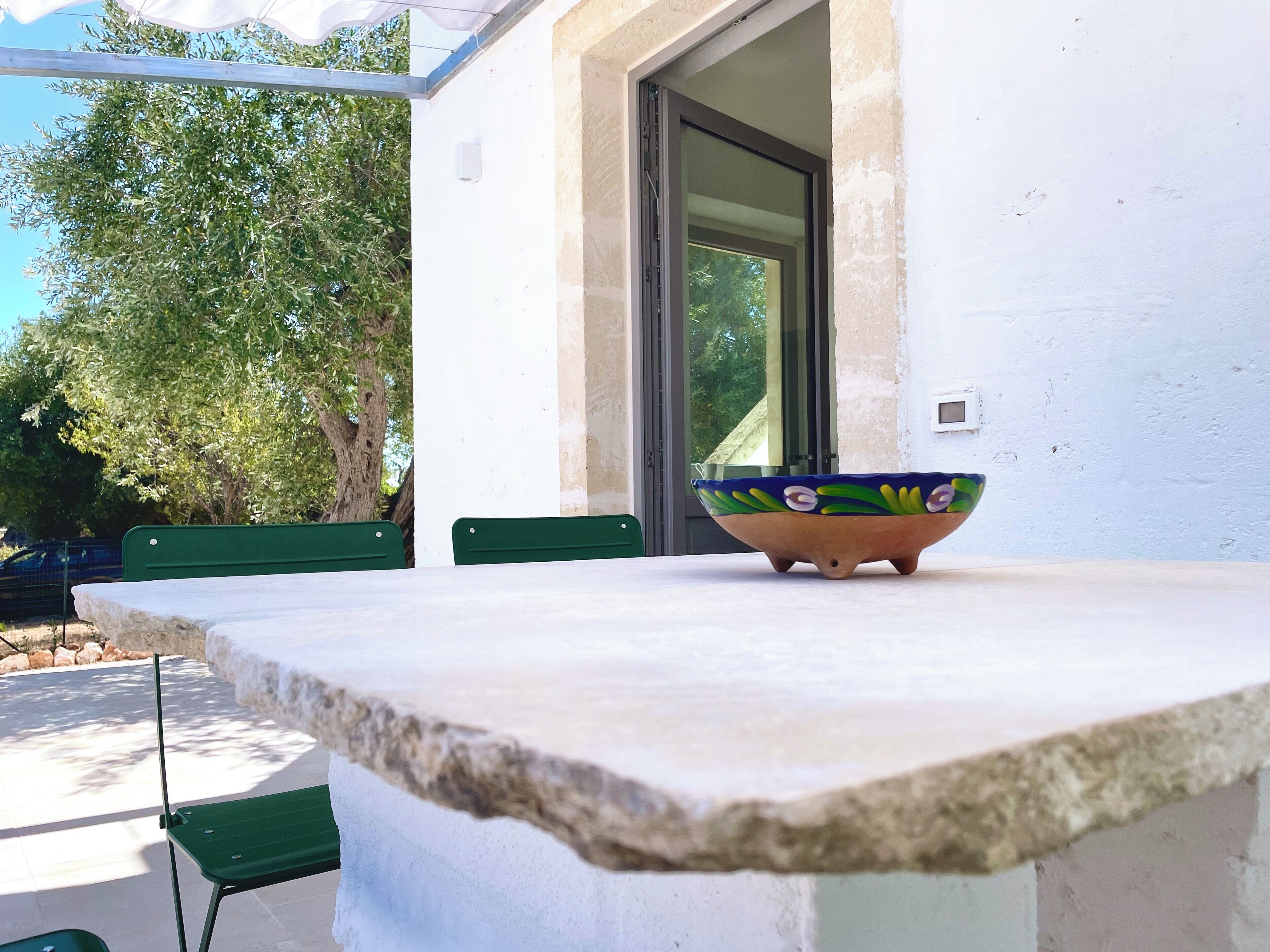 A table with a colorful bowl, green chairs, and a white wall with a window.