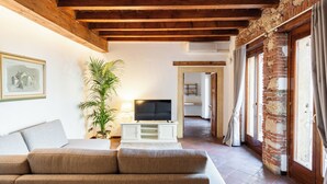 A living room with a sofa, a TV on a white stand, a potted plant, and a brick wall. - Residenza Merici - Elegante e Fiabesca (Verona)