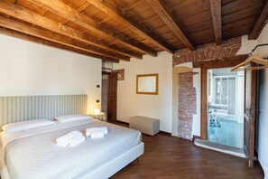 A bedroom with a large bed, wooden beams, a brick wall, and a bathroom visible through an open door. - Residenza Merici - Elegante e Fiabesca (Verona)