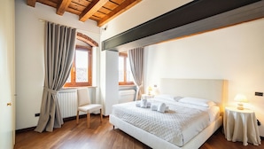 A bedroom with a large bed, wooden flooring, a bench, a bedside table with a lamp, and curtains on the windows. - Residenza Merici - Elegante e Fiabesca (Verona)