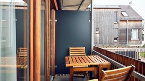En balkon med et træbord og stole, en mørk væg og udsigt til omkringliggende bygninger.