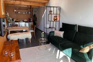 Een gezellige woonkamer met een groene bank, een houten salontafel en een klein bijzettafeltje. Op de achtergrond een eethoek met een tafel en stoelen.