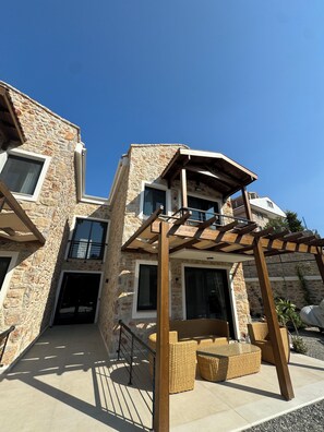 A multi-story stone building with a covered outdoor seating area and a pergola. - ULTAVA KORU SUITES (Datca)