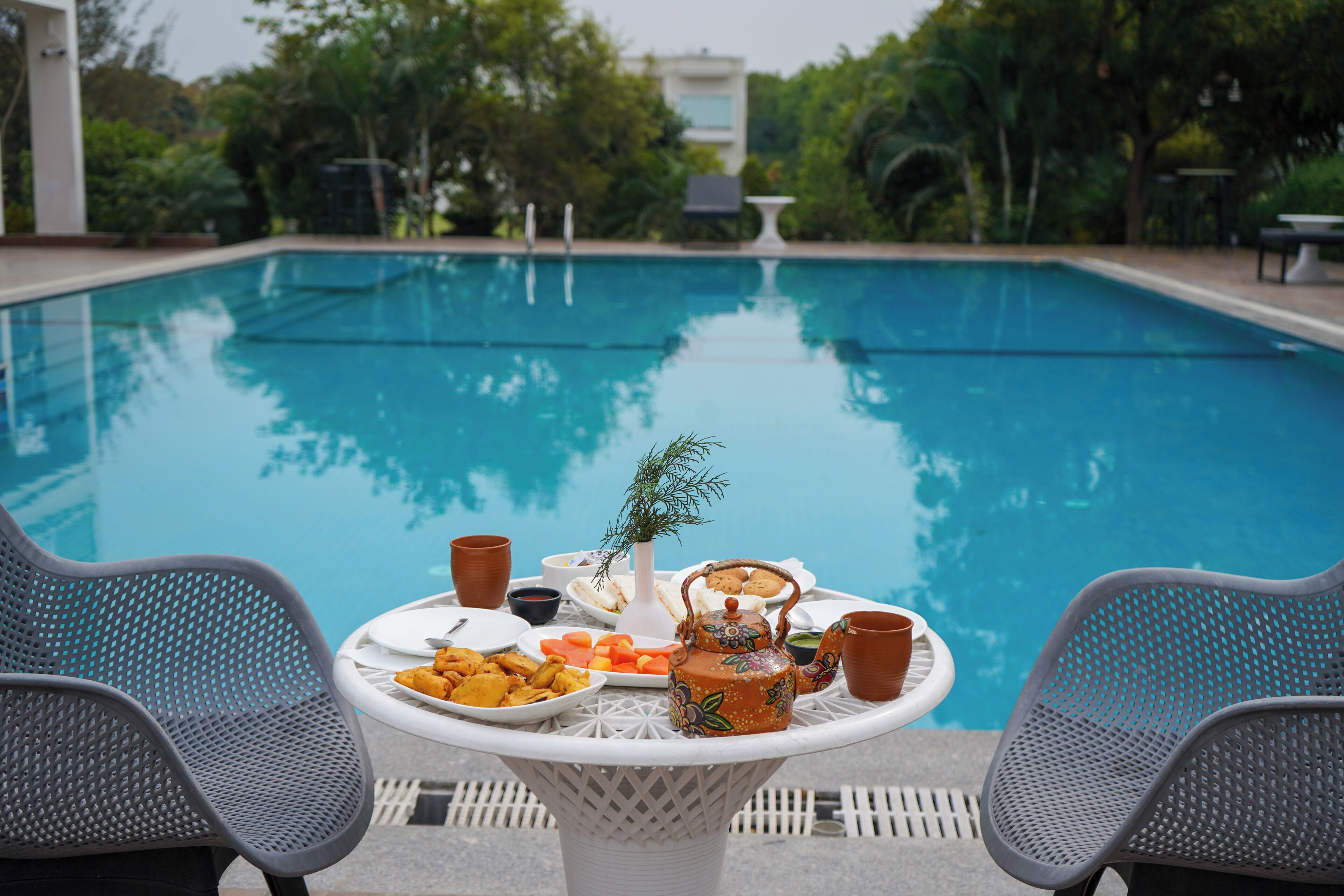Una mesa junto a la piscina con comida y bebidas, rodeada de sillas, y una piscina al fondo.