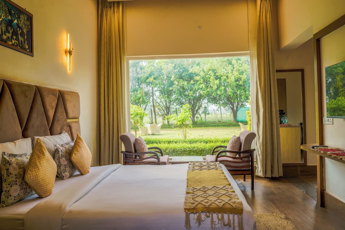 A hotel room with a large bed, two armchairs, a small table, and a view of a garden.