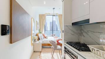 A modern hotel room with a bed, a kitchen area, and a large window.