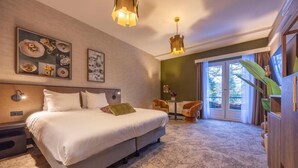 Una habitación de hotel con una cama grande, una mesita con sillas, un ventanal con cortinas y una planta.